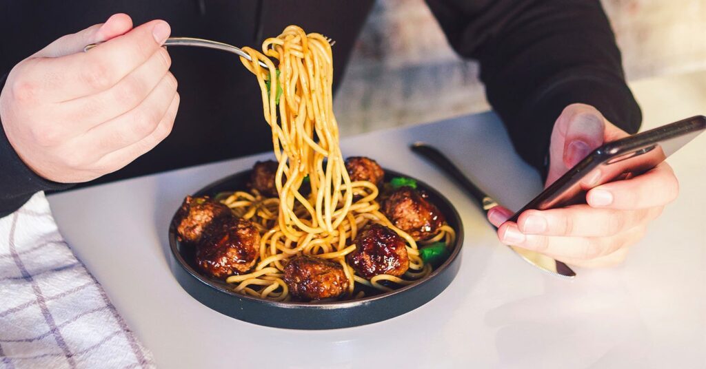Photo shows someone eating a bowl of spaghetti and meatballs while looking at their phone. Who says plant-based food can't be delicious and nutritious.