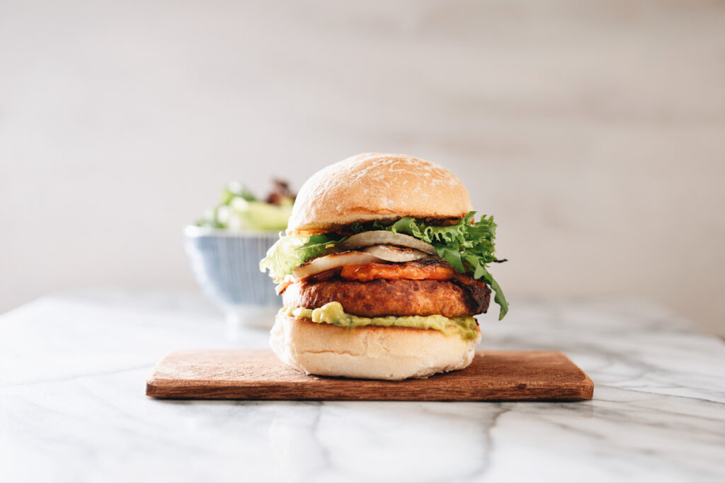 Photo shows a tall vegan burger with salad, quacamole, and sauce on a wooden board. Who says plant-based food can't be delicious and nutritious.