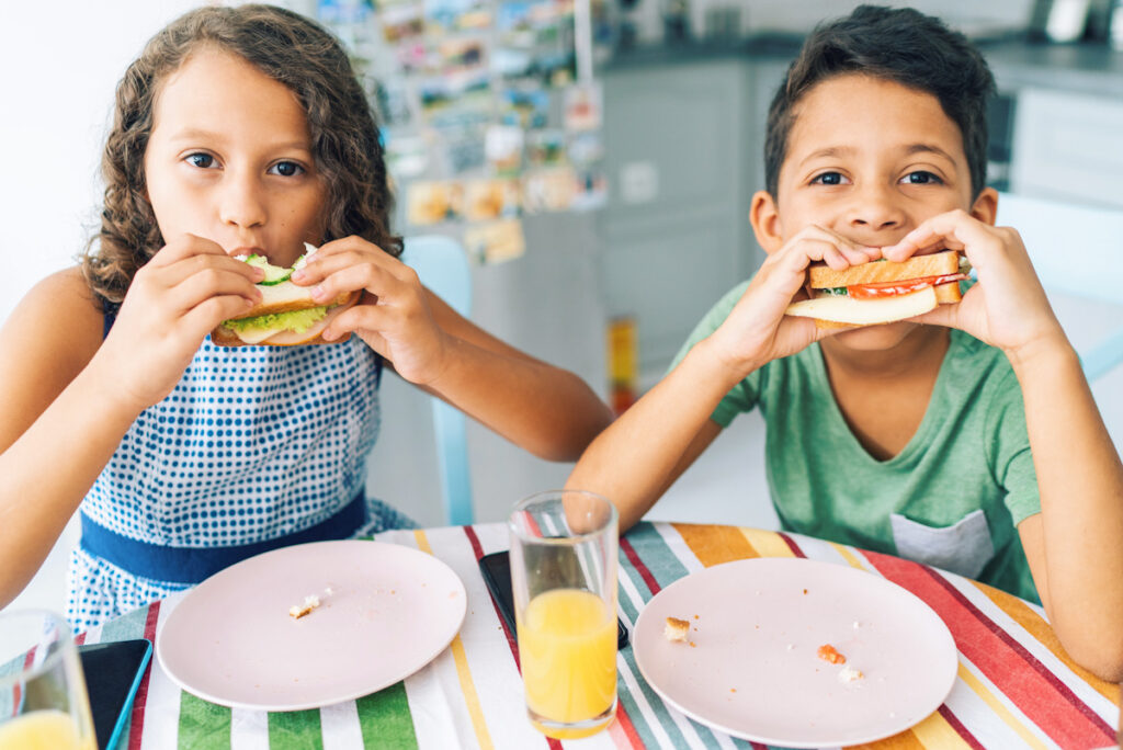 Photo shows two children eating salad-filled sandwiches at a dining table.