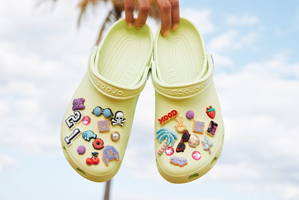 Photo of someone holding a pair of sticker-covered, pale yellow vegan crocs up by the heels against a sky and palm tree background.