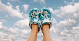 Photo of someone's feet wearing bright blue and white, tie-dyle style vegan crocs, feet held up against a sky background.