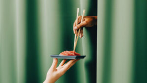 What is a cultured sushi tasting room? Image shows a hand holding up a plate of sushi with another hand emerging from a green curtain with chopsticks.