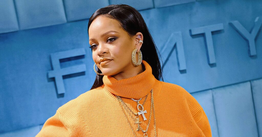Rihanna in an orange jumper in front of a blue Fenty sign