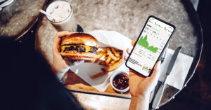 Photo shows someone eating a burger and fries with their phone in one hand, showing a graph—a plant-based market report predicts continued growth in the coming years.