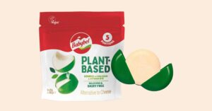 Photo shows a bag of plant-based Babybel cheese on a light orange background. A cheese wheel in green wax sits next to the bag.