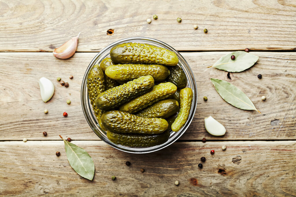 Photo features a bowl of pickled gherkins, surrounded by whole peppercorns, bay leaves, and garlic cloves.