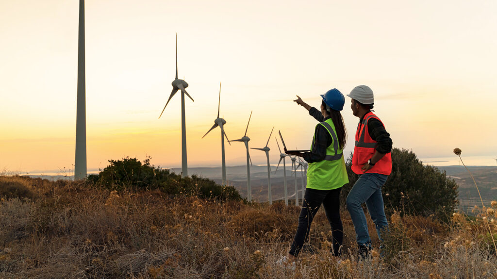 Photo shows two workers in hi-vis jackets and helmets pointing at a row of wind turbines. A new Illinois law plans to phase out fossil fuels in favor of clean energy such as wind.