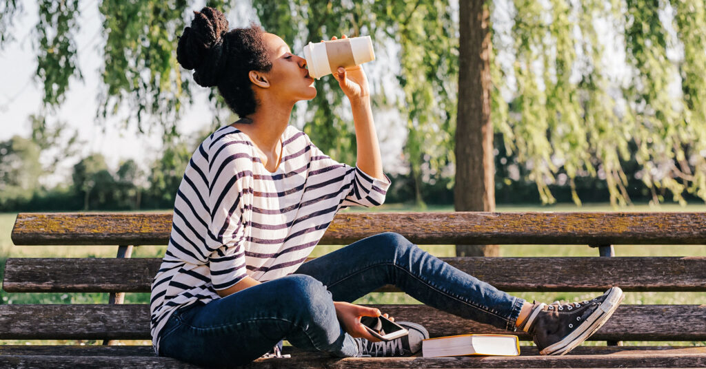 Photo shows a woman sat on a park bench drinking coffee from a takeout cup. Here are 5 caffeine-free alternatives to coffee.