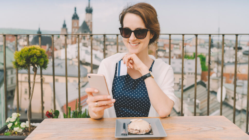 Photo shows a woman wearing sunglasses eating on a rooftop and using her phone. Google has just made it possible to search for hotels by sustainability rating.