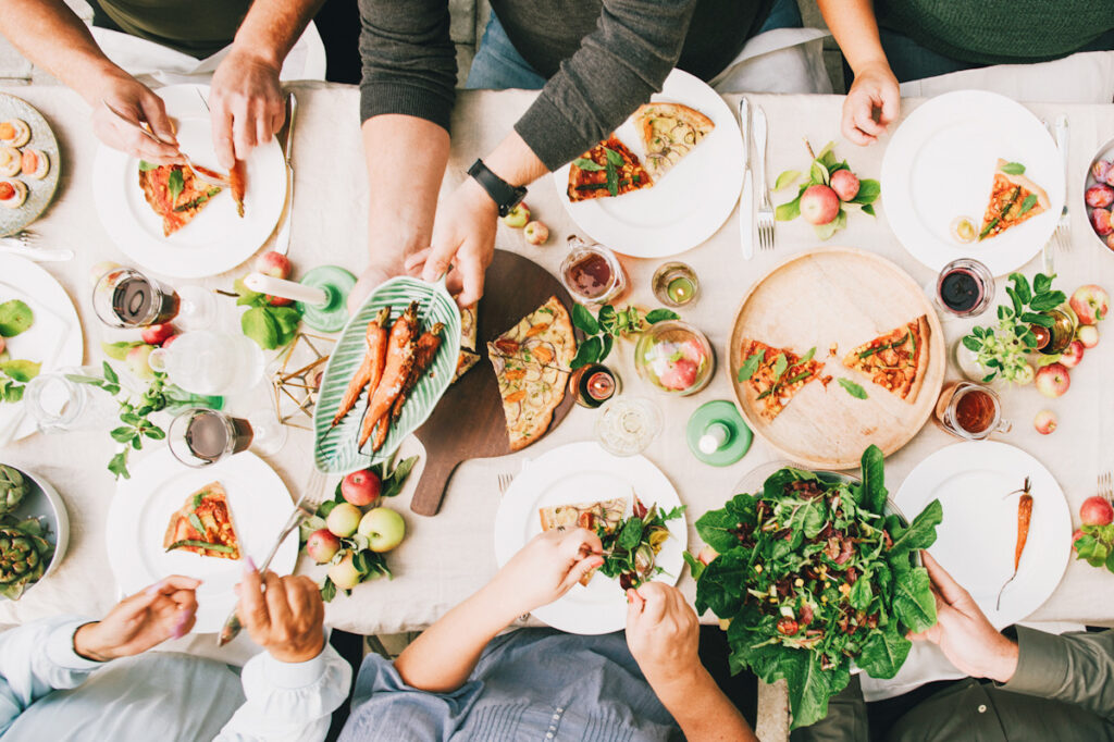 Photo is an overhead shot of a table with many people sat sharing food and passing plates.