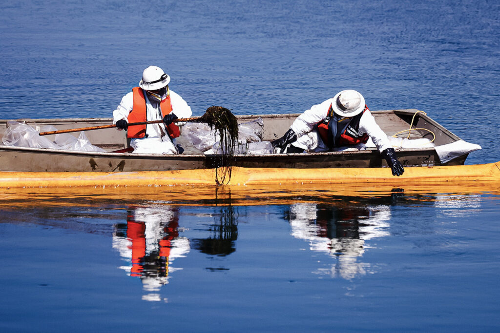 Photo shows two people in protective gear cleaning the oily water.