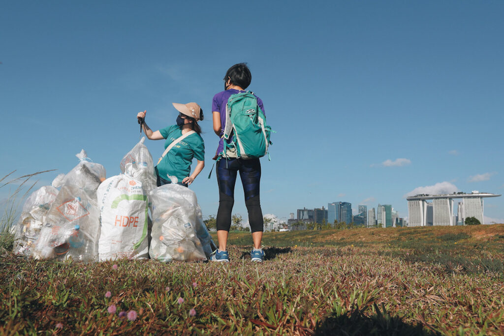 Photo shows two people collecting recyclables in large bags in an outdoor setting.