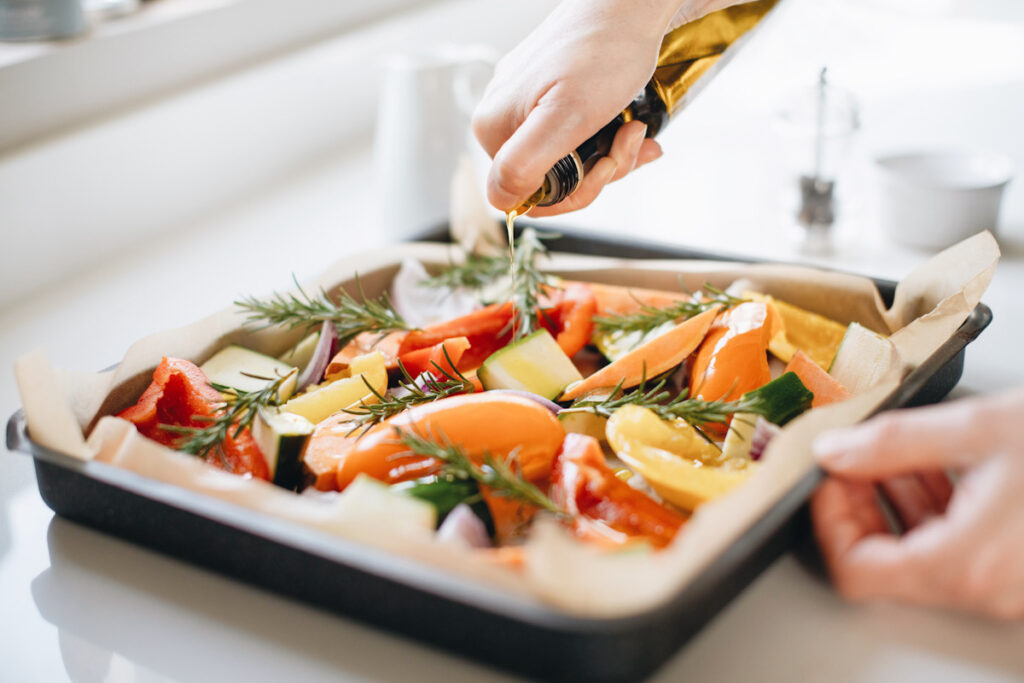 Photo shows someone pouring olive oil over a sheet pan full of vegetables and seasoning, ready to go in the oven.