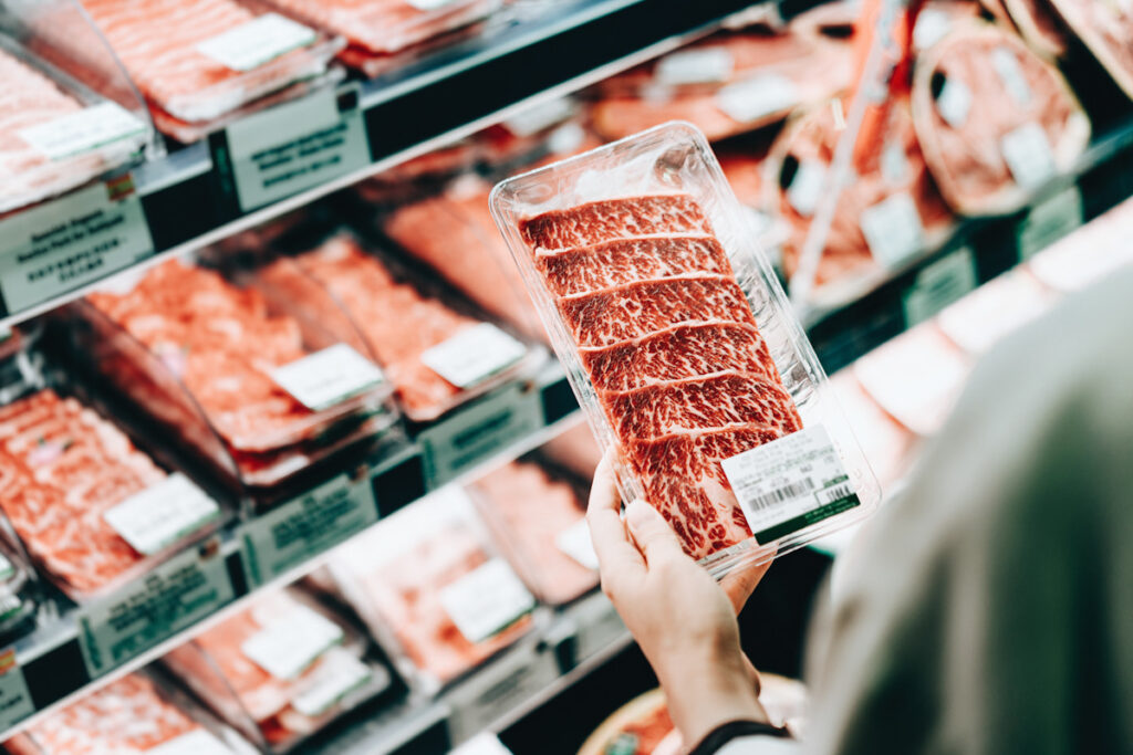 Over the shoulder view of young Asian woman shopping in a supermarket. She is choosing meat and holding a packet of organic beef in front of the refrigerated section Processed with VSCO with a6 preset