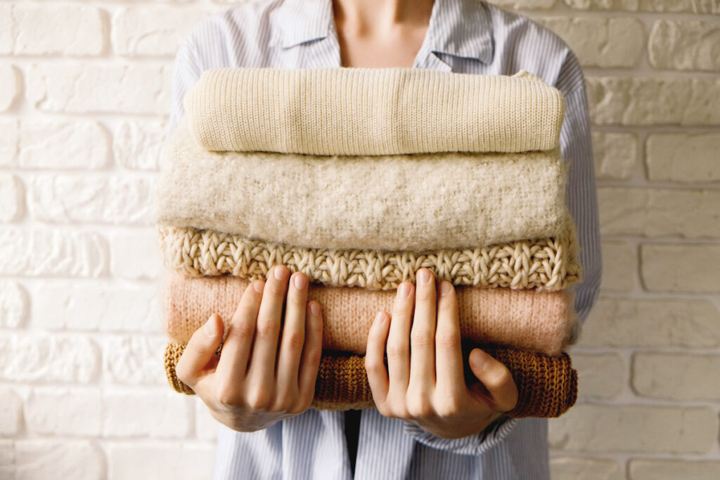 Photo shows a person holding a stack of folded clothes to her chest, including wool-like sweaters.
