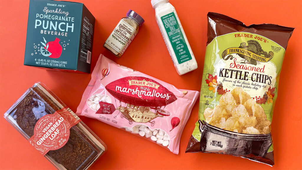 Photo shows a selection of the vegan-friendly items new at Trader Joe's for November 2021 on an orange background, including gingerbread, chips, and marshmallows.