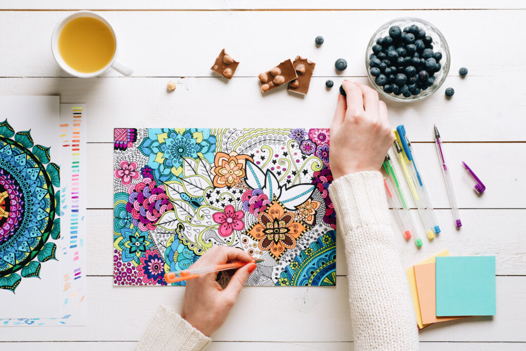 Photo shows a pair of hands drawing in an adult coloring book next to a cup of tea, blueberries, and chocolate.