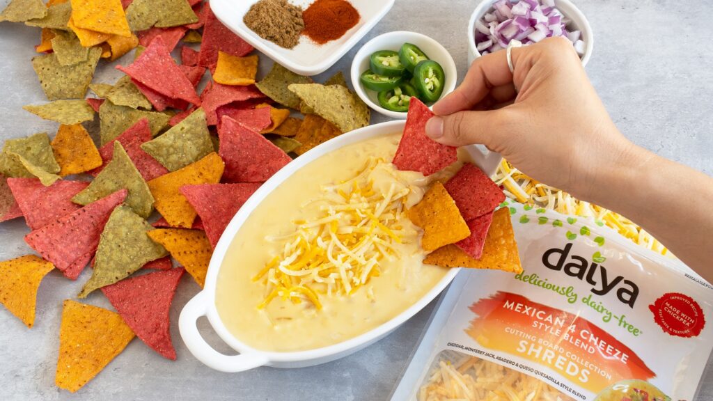 Photo shows vegan queso in a ceramic dish with a side of colorful tortilla chips. A bag of Daiya Mexican cheese shreds is on the side.