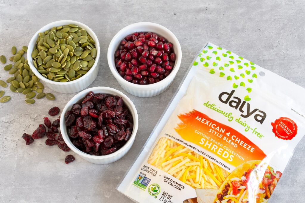 Photo shows Daiya vegan cheese shreds with three ramekins filled with pepitas, pomegranate arils, and dried cranberries, respetively.