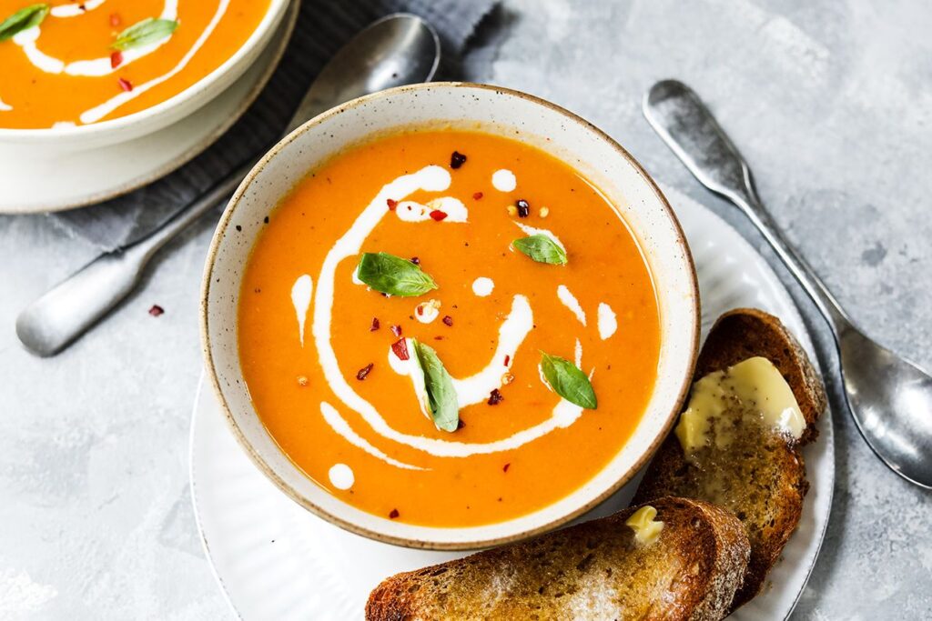 Image shows a bowl of tomato soup topped with a swirl of coconut milk, chili flakes, and fresh basil