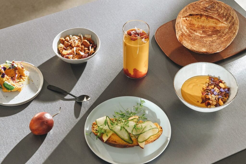 Photo shows a table with a bowl of granola, a smoothie, a sourdough boule, and toast