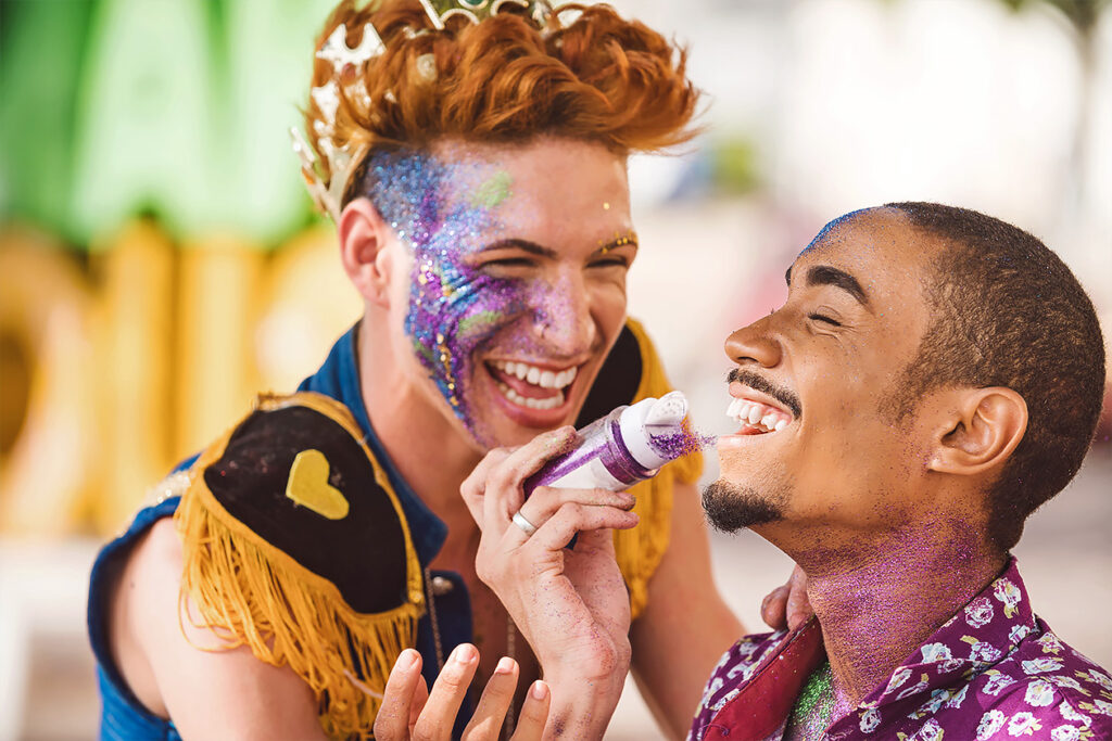 Photo shows two men applying glitter to each others faces and laughing together.