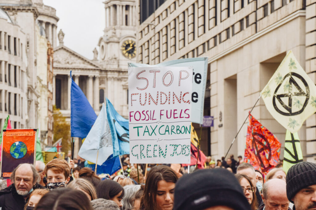 Photo shows Extinction Rebellion protestors marching. one person holds a sign that reads "stop funding fossil fuels; tax carbon; green jobs."