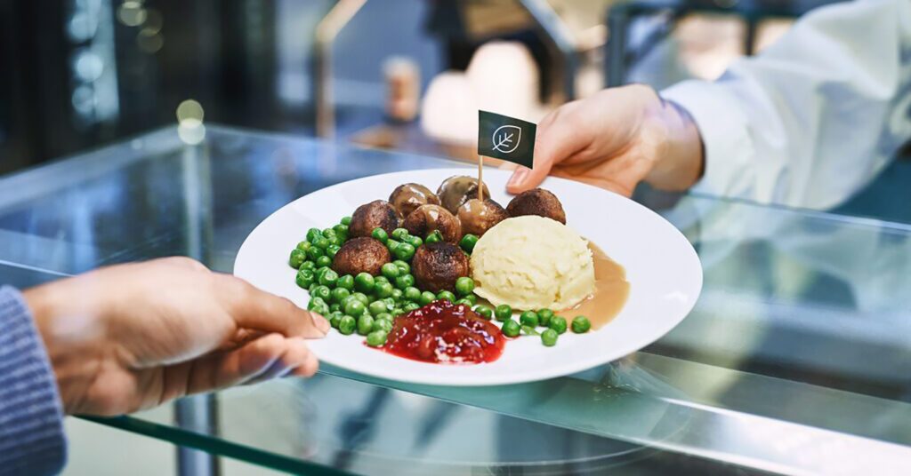 Photo shows a person handing someone else a plate of plant-based meatballs, mash, vegetables, and chutney. IKEA is 3D-printing vegan meatballs.