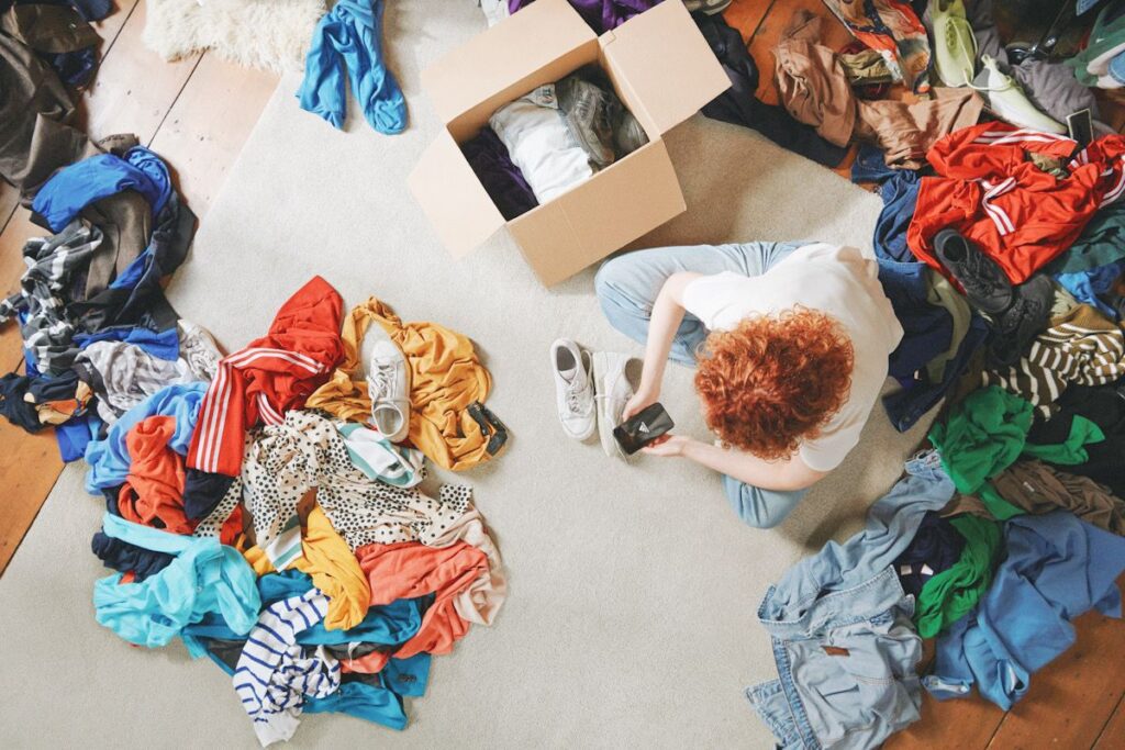 A person sits on the floor surrounded by clothes and boxes