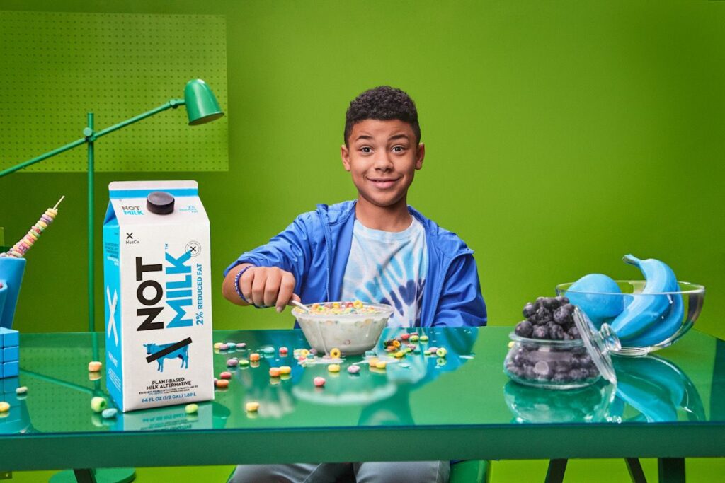 Photo shows a kid in a blue windbreaker eating a bowl of Froot Loops with a carton of NotMilk, a vegan milk product.